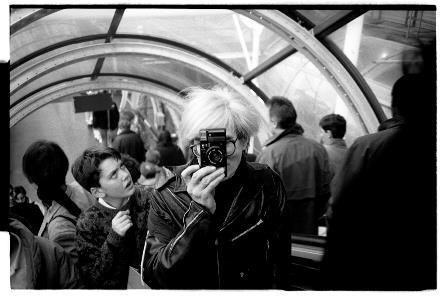Andy Warhol sosteniendo una cámara frente a su rostro. Andy Warhol biografía 